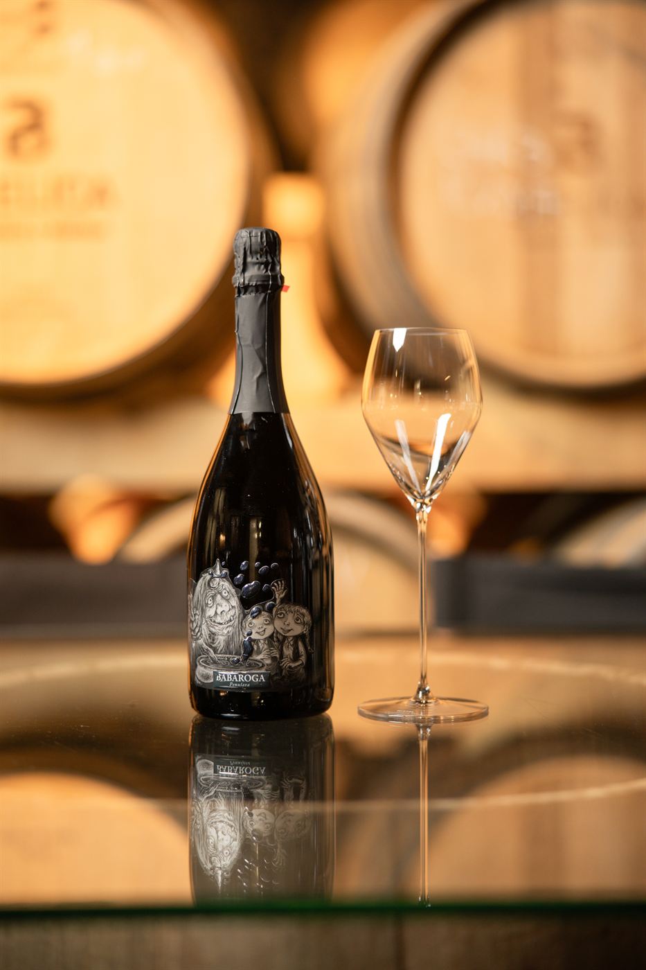 A bottle of Sparkling Babaroga with a glass in front of barriques